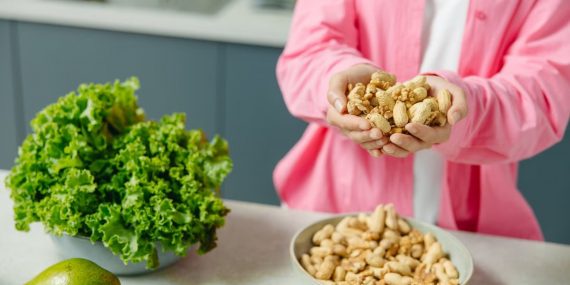 Hands presenting peanuts with a bowl, lettuce, avocado, and tomato nearby on kitchen counter.